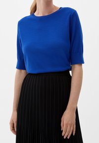 Kvinna i en kungligt blå kortärmad tröja instoppad i en hög midja svart plisserad kjol, stående mot en helt vit bakgrund.