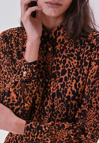 Chemise à imprimé léopard orange et noir, avec un col, des boutons dorés et un tissu texturé avec une coupe ample.