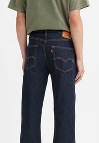 Dark blue denim jeans with a relaxed fit, visible yellow stitching, and two back pockets featuring a curved design. Leather brand patch attached.