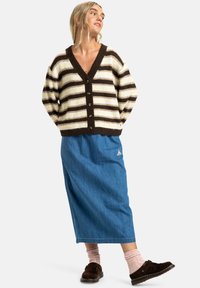 Gestreepte cardigan in crème en bruin, met een diepe V-hals en knopen. Gecombineerd met een blauwe spijkerrok en bruine instappers.