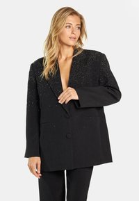 Blazer negro con un profundo escote en V, corte oversized y adornado con pequeños acentos brillantes en los hombros. Cierre de doble botón.