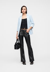 Γυναίκα που φοράει ανοιχτό μπλε σακάκι, μαύρο πουκάμισο με κουμπιά, μαύρο δερμάτινο παντελόνι, κρατώντας μαύρη τσάντα, στέκεται μπροστά σε λευκό φόντο.