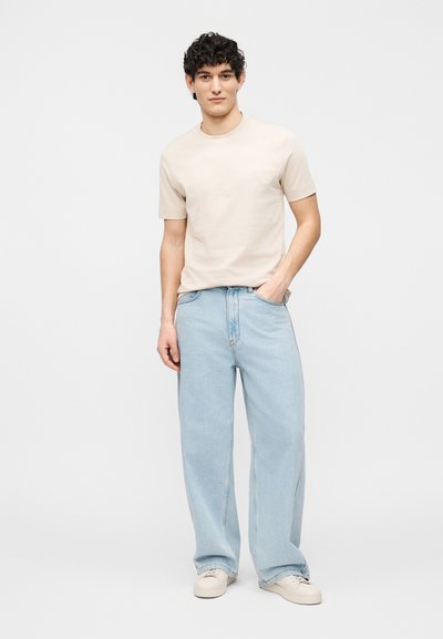 Νέος άνδρας με κατσαρά μαλλιά, φορώντας μπεζ κοντομάνικο πουκάμισο, ανοιχτό μπλε παντελόνι με φαρδιά μπατζάκια και λευκά αθλητικά παπούτσια, στέκεται με το ένα χέρι στην τσέπη.