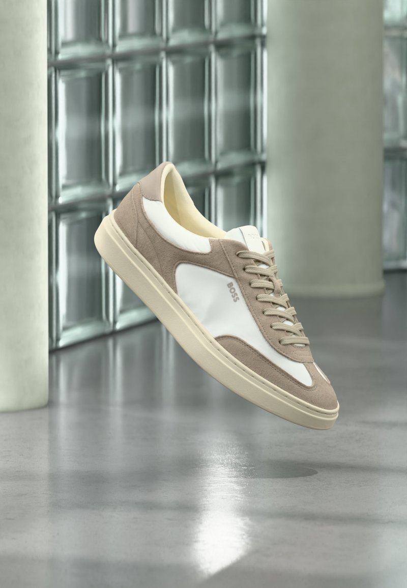 Baskets en suède beige et blanc avec lacets et logo « BOSS », flottant au-dessus d’un sol en béton poli avec des murs en blocs de verre en arrière-plan.