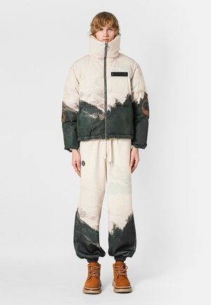 Chaqueta acolchada color crema con estampado de montañas en verde oscuro, cierre con cremallera, cuello alto; combinada con pantalones claros, botas marrones y detalle de logotipo circular.