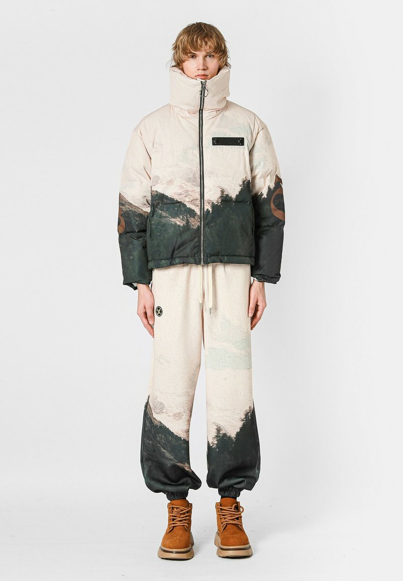 Chaqueta acolchada color crema con estampado de montañas en verde oscuro, cierre con cremallera, cuello alto; combinada con pantalones claros, botas marrones y detalle de logotipo circular.