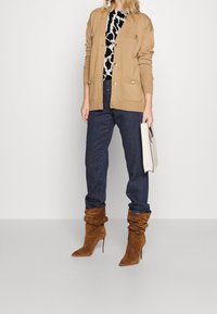Beige cardigan with gold buttons, black and white patterned top, dark jeans, and brown slouchy ankle boots. Holding a light beige clutch.