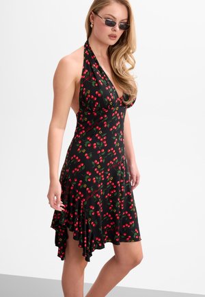 Frau mit schwarzem Neckholder-Kleid mit rotem Kirschmuster, asymmetrischem Saum, Sonnenbrille und welligem blondem Haar steht vor neutralem Hintergrund.