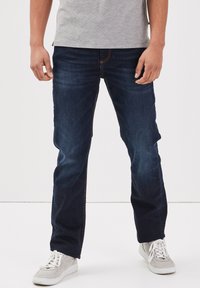 Jeans de mezclilla azul oscuro con corte recto, que presentan costuras naranjas, una cremallera estándar y se llevan con zapatillas blancas.