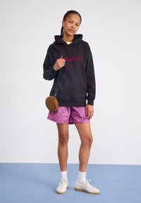 Black hoodie with red logo, pink leopard print shorts, white socks, and beige sneakers. Brown circular bag with strap. Simple background.