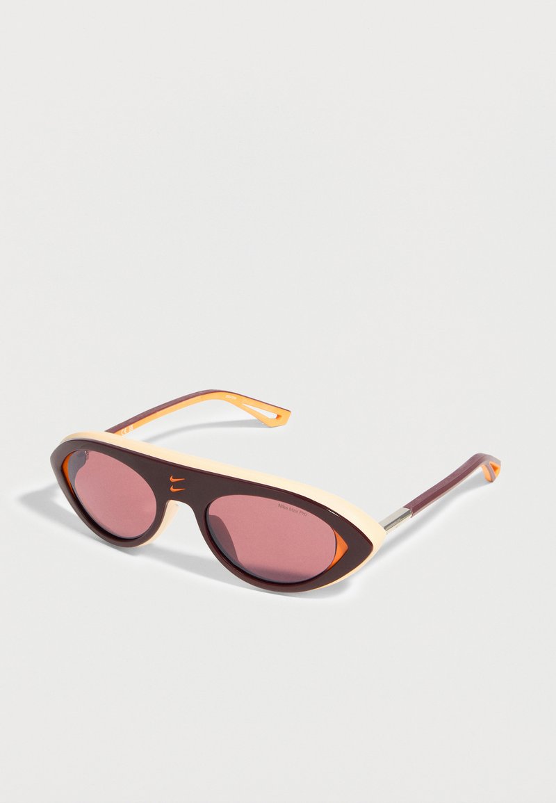 Gafas de sol con montura estilo ojo de gato en marrón oscuro y crema, lentes tintadas de rosa y detalles en naranja, dispuestas en ángulo sobre un fondo blanco.