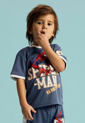Jeune enfant aux cheveux bruns portant un t-shirt et un short Spider-Man bleus, tenant un doigt près de la bouche sur un fond bleu clair.