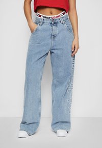 Světlé modré džínové kalhoty s širokými nohavicemi, vyrobené z denimu, s bílým potiskem loga podél levé strany a značkovým páskem.