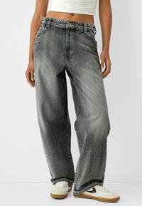 Osoba ubrana w szerokie, wyblakłe czarne dżinsy, białą krótko skrojoną bluzkę i białe sneakersy z gumową podeszwą.