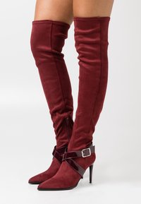 Burgundy knähöga stövlar i mocka, med spetsiga tår, hög klack och ett glänsande vristband med spänne.