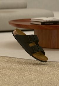 Μαύρο σανδάλι με δύο ρυθμιζόμενες αγκράφες, πάτο από φελλό και υφασμένο λαστιχένιο πέλμα, αιωρούμενο πάνω από ένα ανοιχτόχρωμο χαλί.