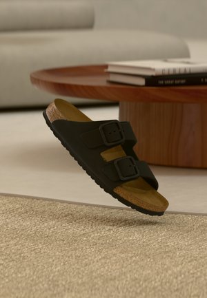 Μαύρο σανδάλι με δύο ρυθμιζόμενες αγκράφες, πάτο από φελλό και υφασμένο λαστιχένιο πέλμα, αιωρούμενο πάνω από ένα ανοιχτόχρωμο χαλί.