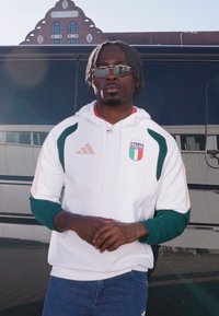 Hanorac alb cu accente verzi și dungi aurii, cu logo-ul echipei naționale a Italiei. Purtat cu blugi albaștri. Păr întunecat, texturat.