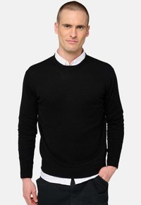 Schwarzer Strickpullover mit Rundhalsausschnitt, langen Ärmeln und glatter Textur, über einem weißen Kragenhemd getragen.