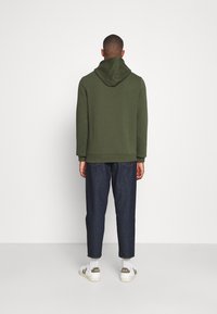 Felpa verde scuro con cappuccio, vestibilità ampia e orlo a costine, abbinata a jeans blu scuro a gamba affusolata e sneakers bianche con dettagli verdi.