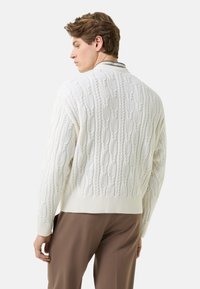 Weißer Strickpullover mit Zopfmuster, einem gerippten Saum und Bündchen sowie einem strukturierten Muster. Das Modell trägt braune Hosen und ist von hinten zu sehen.
