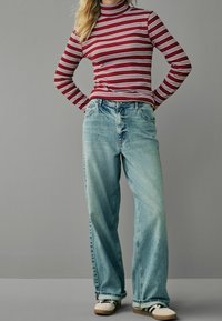 Personne portant un col roulé rayé rouge et blanc, un jean large bleu clair, et des baskets blanches avec des rayures noires, se tenant les mains sur les hanches.