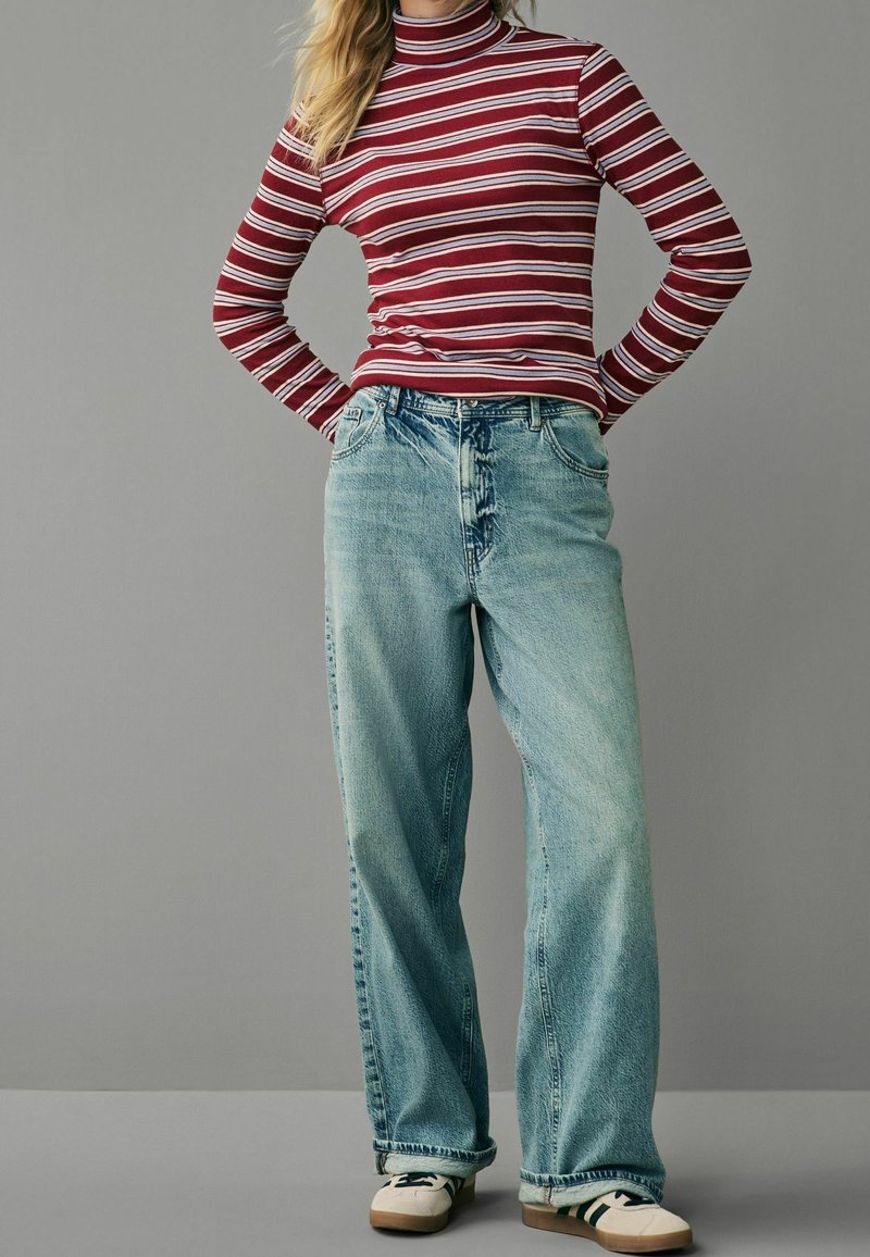 Personne portant un col roulé rayé rouge et blanc, un jean large bleu clair, et des baskets blanches avec des rayures noires, se tenant les mains sur les hanches.