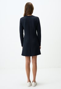 Robe longue à manches longues de couleur marine avec une silhouette évasée, un tissu texturé et un détail de couture à l'arrière. Portée avec des baskets blanches.