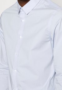 Chemise boutonnée bleu clair en tissu lisse, dotée d'un col classique, de boutons blancs et d'un logo "OK" discret sur la poitrine gauche.