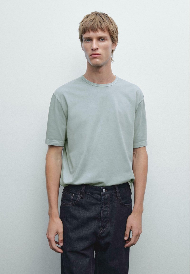 Light green cotton t-shirt with a round neck and short sleeves, paired with dark denim jeans, standing against a neutral background.