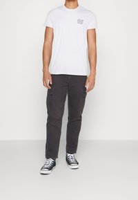 Camiseta blanca de manga corta con texto impreso en el pecho, combinada con pantalones cargo grises oscuros con bolsillos de parche y zapatillas negras.