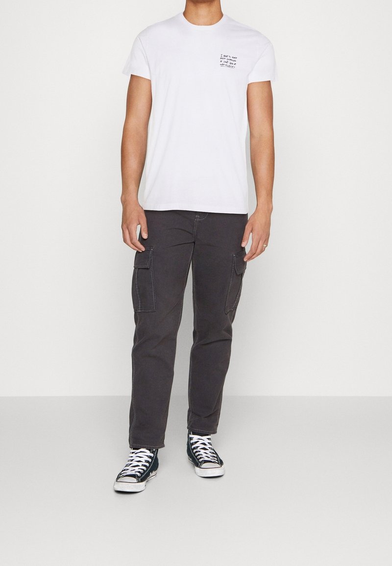 Camiseta blanca de manga corta con texto impreso en el pecho, combinada con pantalones cargo grises oscuros con bolsillos de parche y zapatillas negras.