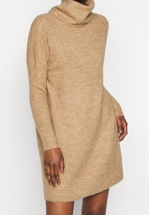 Langærmet beige strikket turtleneck kjole båret af en person med hænderne afslappet ved siden mod en ensfarvet baggrund.
