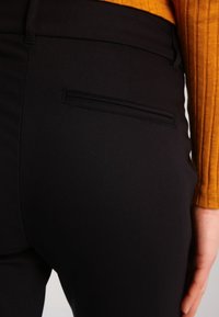 Närbild av en svart byxficka på höger höft bärande med en senapsgul, randig långärmad topp och en synlig hand vid sidan.