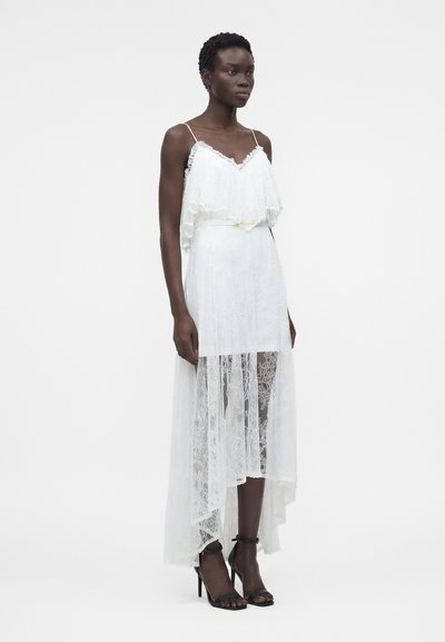 Γυναίκα με λευκό δαντελωτό φόρεμα high-low με λεπτές τιράντες, ζώνη στη μέση και μαύρα ψηλά τακούνια με λουράκι στον αστράγαλο, στέκεται μπροστά σε λευκό φόντο.