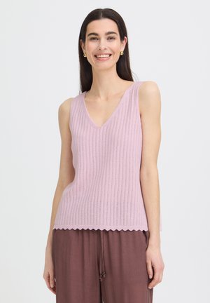 Donna che indossa un top lavorato a maglia senza maniche di colore viola chiaro con orlo smerlato e pantaloni marroni larghi, sorridente davanti a uno sfondo semplice.