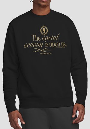 Man wearing black sweatshirt with gold text: "The social season is upon us. Lady Whistledown Bridgerton" and a decorative silhouette above text.