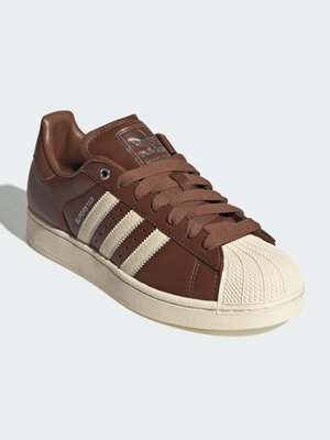 Brun Adidas Superstar sneaker med creme farvet skal tå, tre creme farvede striber på siden og brune snørebånd på hvid baggrund.