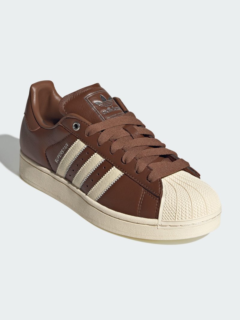 Καφέ αθλητικό παπούτσι Adidas Superstar με κρεμ χρώματος μύτη, τρεις κρεμ ρίγες στο πλευρό και καφέ κορδόνια σε λευκό φόντο.