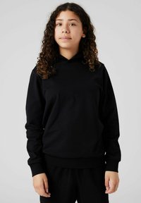 Junge Person mit lockigem Haar, die einen schwarzen Hoodie und passende schwarze Hosen trägt und vor einem einfachen hellen Hintergrund steht.