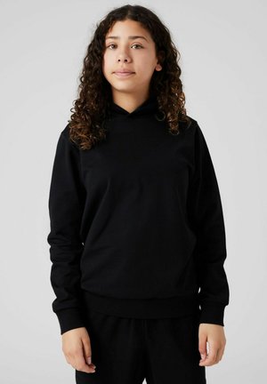 Jong persoon met krullend haar, gekleed in een zwarte hoodie en bijpassende zwarte broek, staande tegen een effen lichte achtergrond.