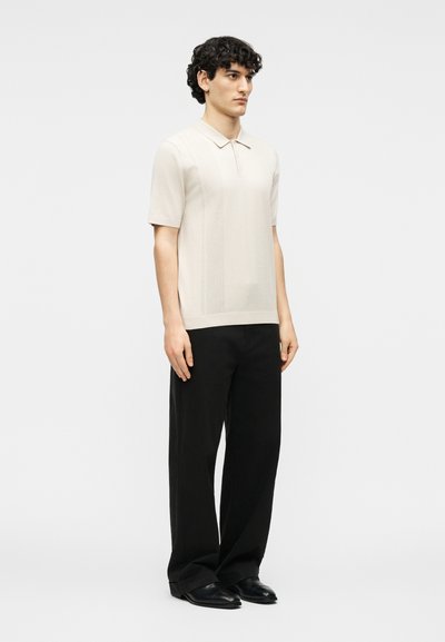 Νέος άνδρας με κατσαρά μαλλιά φοράει μπέζ κοντομάνικη πόλο μπλούζα, μαύρο παντελόνι φαρδύ, και μαύρα δερμάτινα παπούτσια, στέκεται ακίνητος.