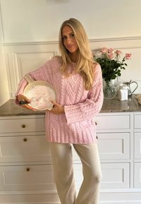 Gaiši rozā adīta džemperis ar kabeli rakumiem, tur rokās maisīšanas bļodu ar balto maisījumu un putotāju. Mīkstas beigu bikses, virtuves fons.