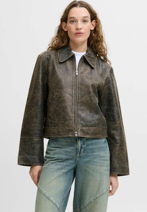 Woman wearing a distressed brown leather zip-up jacket with wide sleeves and light blue, high-waisted jeans.