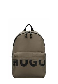 Mochila de tela beige con un acabado texturizado, que presenta un gran logotipo negro "HUGO", doble cremallera, bolsillo frontal y un asa superior.