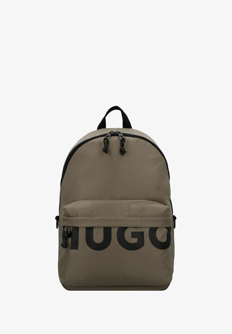 Beige stoffen rugzak met een gestructureerd oppervlak, voorzien van een groot zwart "HUGO" logo, dubbele ritsen, een voorvak en een handvat aan de bovenkant.