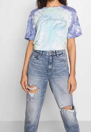 T-shirt bleu clair en tie-dye avec "Los Angeles CA" en gris, associé à un jean bleu clair usé avec de grandes déchirures aux genoux.