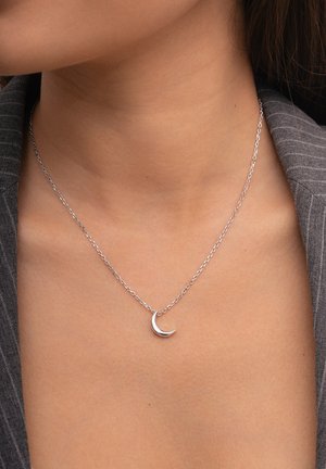 Collana d'argento con un piccolo pendente a forma di mezzaluna su una catena sottile, posizionata su un indumento grigio texturizzato.