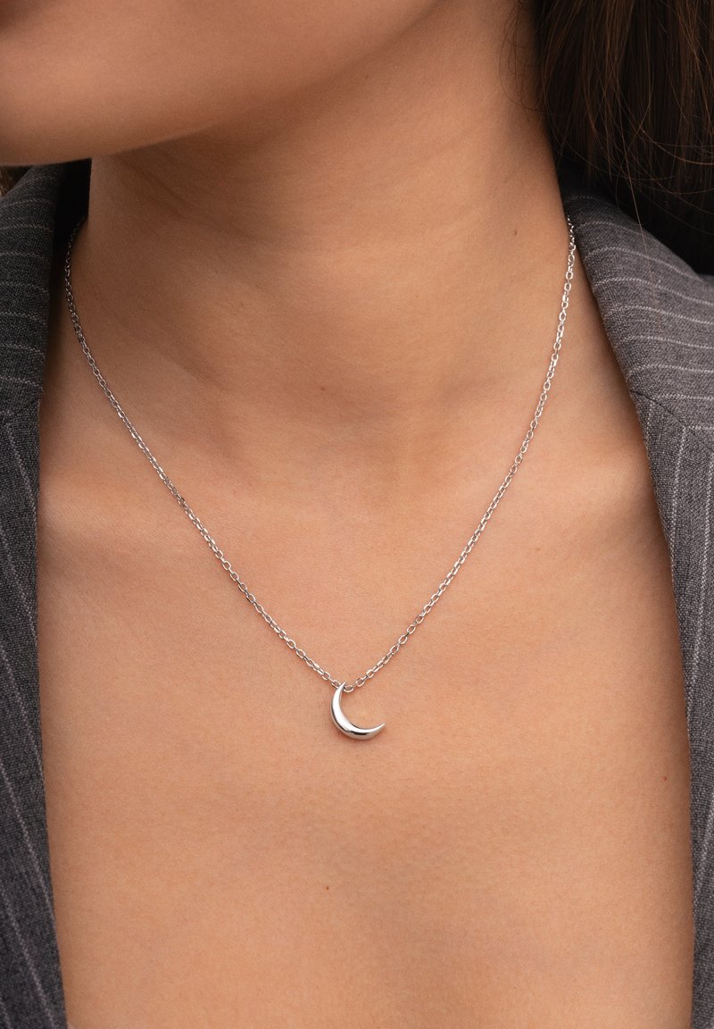 Collar de plata con un pequeño colgante de media luna sobre una cadena delgada, situado sobre una prenda gris texturizada.