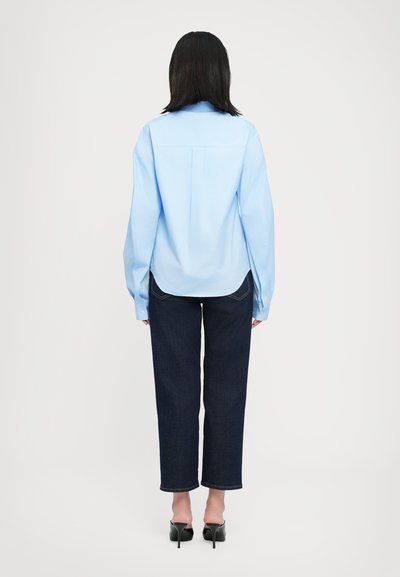 Light blue, long-sleeve shirt with a relaxed fit and curved hem. Paired with dark denim pants and black, pointed-toe heels.